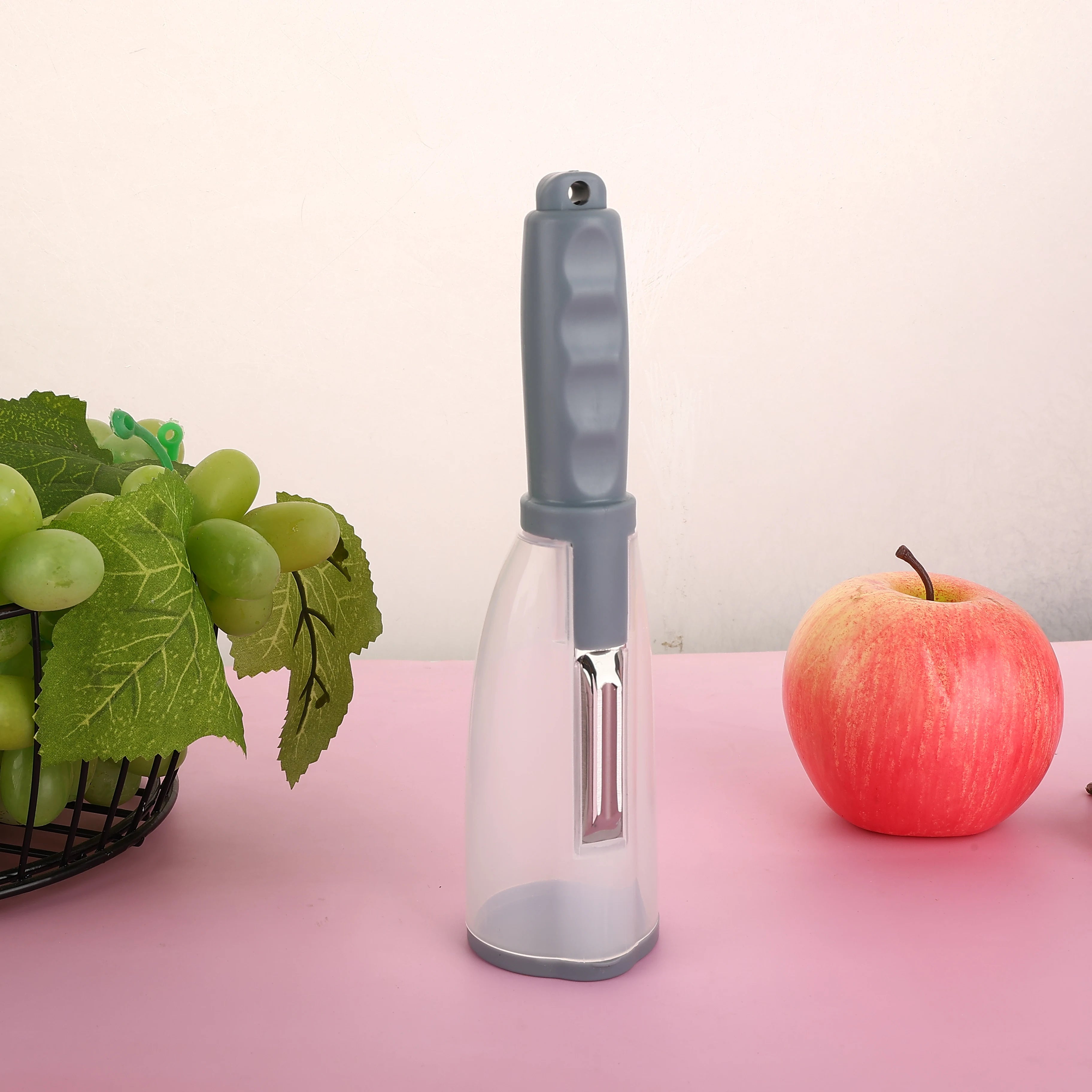 Vegetable Peeler with Container - CozyNest Hub premium handheld tool in green, displayed with fruits on a pink background.